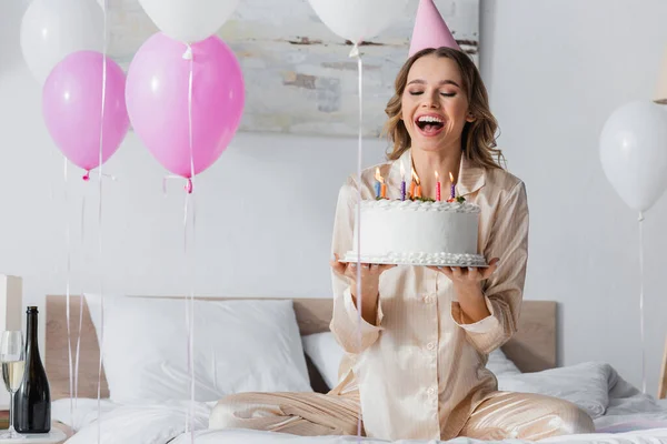 Donna felice in berretto partito tenendo torta di compleanno vicino palloncini e champagne in camera da letto — Foto stock