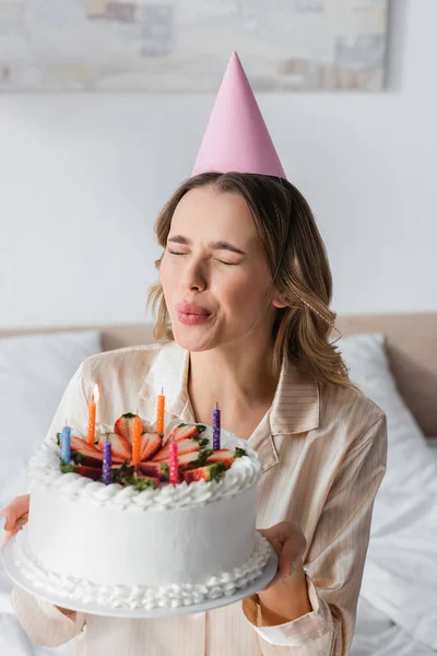 Mujer en pijama y gorra de fiesta soplando velas en pastel de cumpleaños - foto de stock