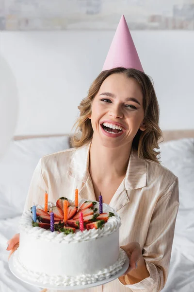 Smiling woman in party cap looking at camera while holding birthday cake at home — Stock Photo