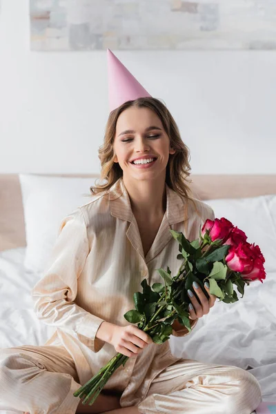 Smiling woman in pajama and party cap holding roses on bed — Stock Photo