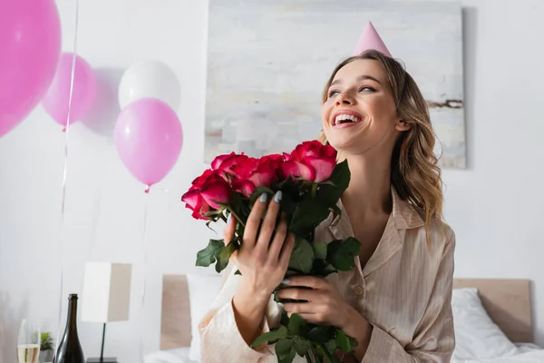 Cheerful woman in party cap holding roses near balloons in bedroom — Stock Photo