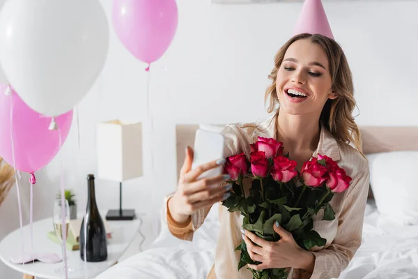 Femme souriante prenant selfie sur smartphone et tenant des roses pendant la célébration de l'anniversaire — Photo de stock