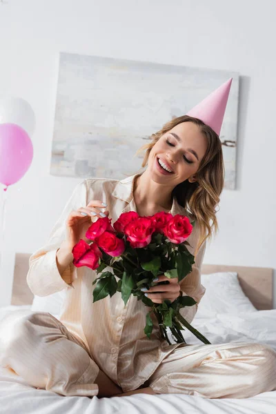 Jovem mulher em pijama e tampão de festa segurando rosas na cama — Fotografia de Stock