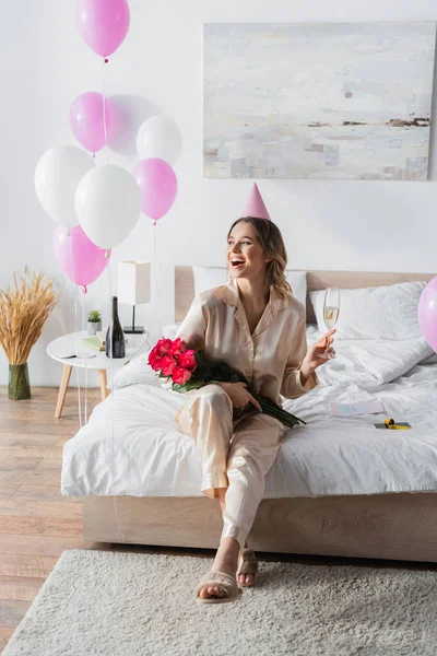 Mujer positiva con gorra de fiesta sosteniendo rosas y champán en la cama - foto de stock