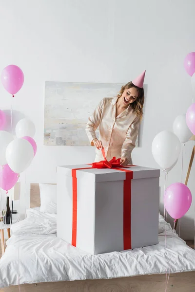 Fröhliche Frau mit Partymütze hält Schleife mit riesigem Geschenk in der Nähe von Luftballons im Schlafzimmer — Stockfoto
