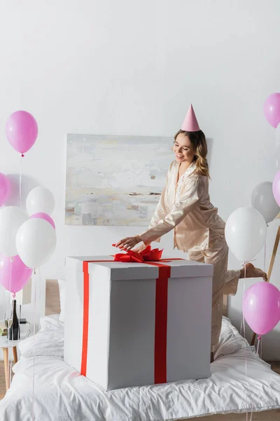 Junge Frau in Pyjama und Partymütze steht neben großem Geschenk auf Bett und Luftballons — Stockfoto