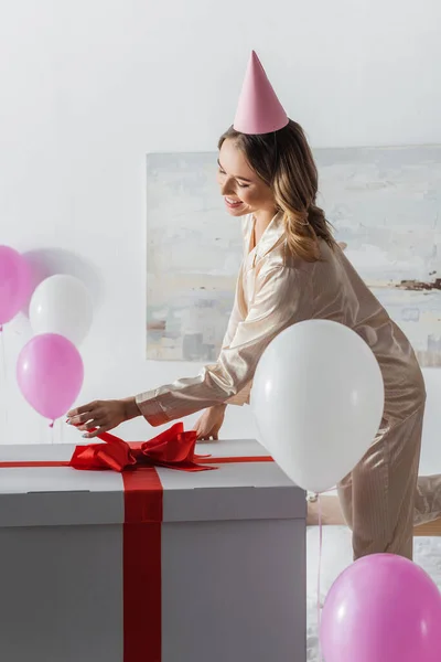 Smiling woman looking at big gift box near balloons in bedroom during birthday celebration — Stock Photo