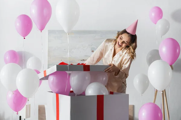 Woman in party cap and pajama opening gift box with festive balloons at home — Stock Photo
