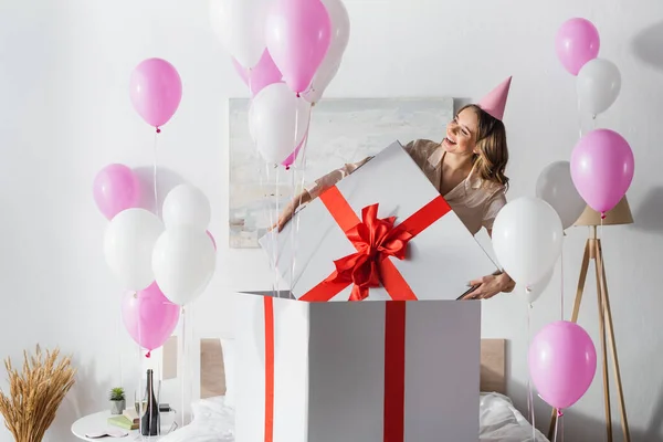 Lächelnde Frau mit Mütze mit riesigem Geschenk neben festlichen Luftballons im Schlafzimmer — Stockfoto