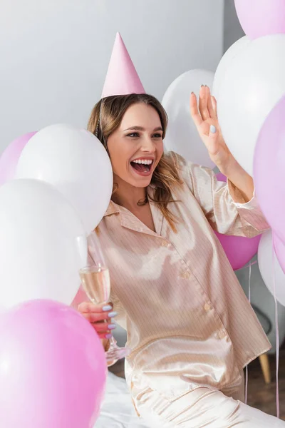 Mulher positiva em boné de festa e pijama segurando champanhe perto de balões borrados em casa — Fotografia de Stock