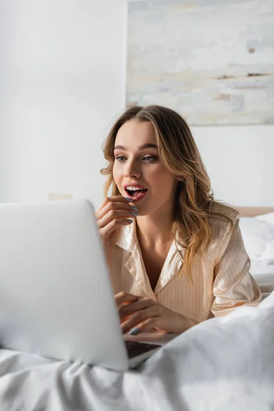 Emocionado freelancer en pijama usando una computadora portátil borrosa en la cama - foto de stock