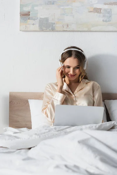 Mujer sonriente en auriculares usando portátil en la cama - foto de stock