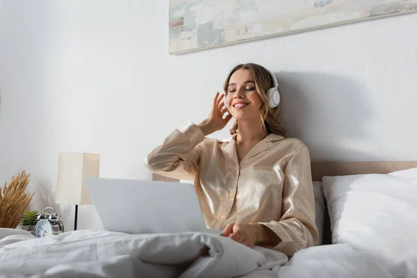 Весела жінка слухає музику в навушниках біля розмитого ноутбука на ліжку — стокове фото