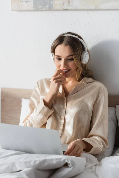 Junge Telearbeiterin mit Kopfhörer und verschwommenem Laptop am Morgen im Bett — Stockfoto