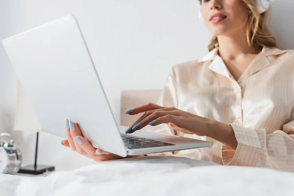 Vista ritagliata della donna sfocata in cuffia utilizzando il computer portatile sul letto — Foto stock