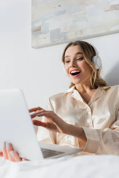 Mulher positiva em fones de ouvido segurando laptop desfocado na cama — Fotografia de Stock