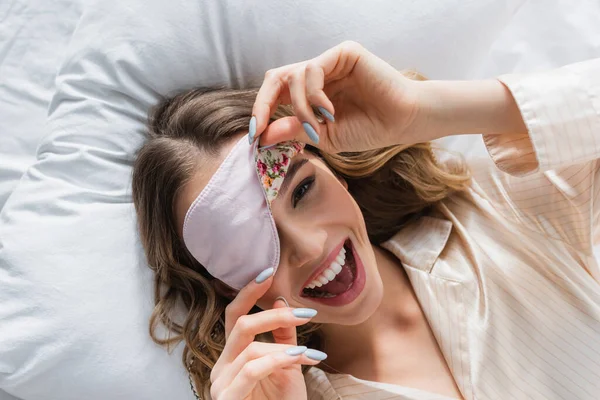Vista superior de la mujer alegre sosteniendo la máscara de dormir en la cama - foto de stock
