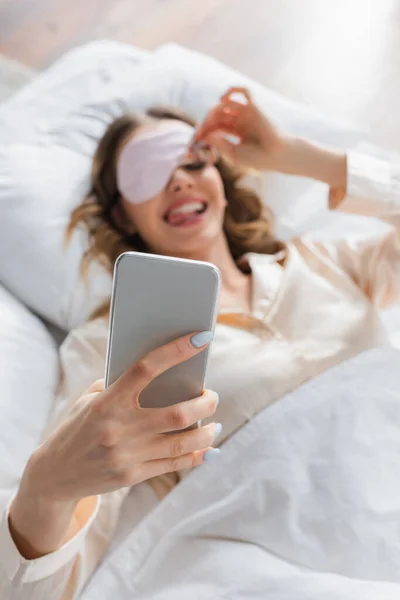 Smartphone en main de femme floue dans le masque de sommeil sur le lit — Photo de stock