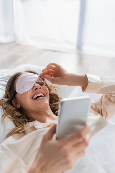 Веселая женщина в маске для сна с помощью смартфона на кровати — стоковое фото