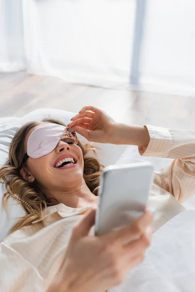 Cheerful woman in sleeping mask using smartphone on bed — Stock Photo
