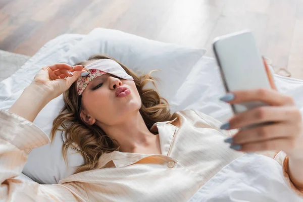 Femme dans le masque de sommeil boudant lèvres et en utilisant smartphone sur le lit — Photo de stock