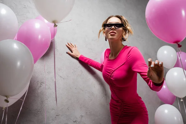Low angle view of stylish woman in sunglasses and dress near festive balloons and concrete wall — Stock Photo
