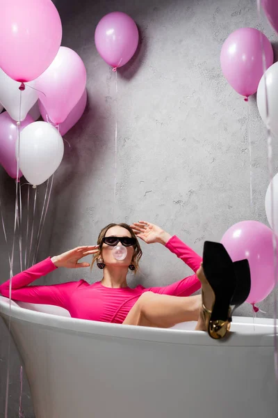 Frau mit Sonnenbrille und Kaugummi sitzt in Badewanne neben festlichen Luftballons — Stockfoto