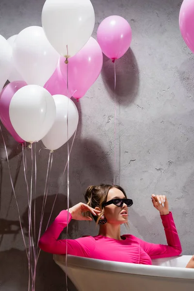Balloons near fashionable woman in sunglasses sitting in bathtub — Stock Photo