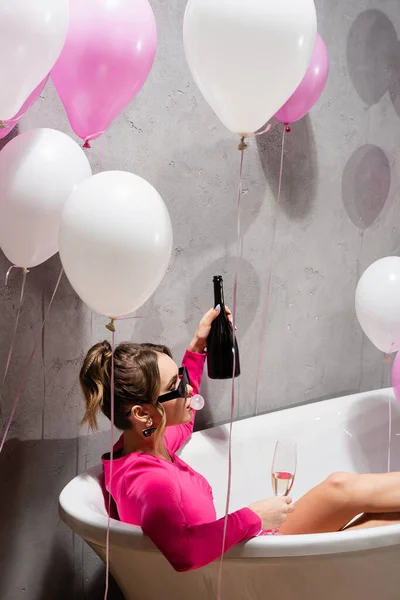 Жінка в сонцезахисних окулярах дме бульбашкою і тримає шампанське у ванній біля повітряних куль — стокове фото