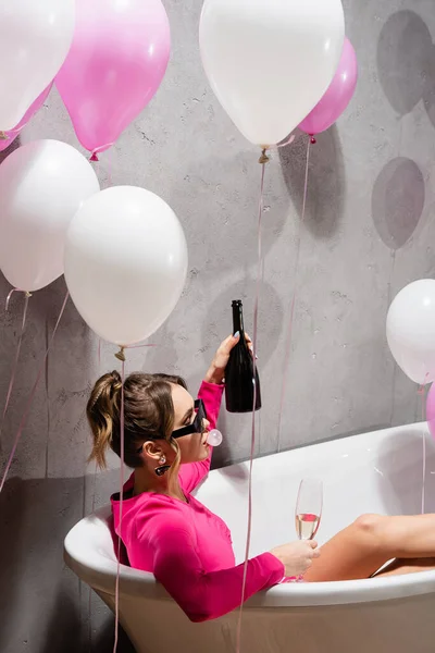 Mujer en gafas de sol soplando goma de mascar y sosteniendo champán en la bañera cerca de globos - foto de stock