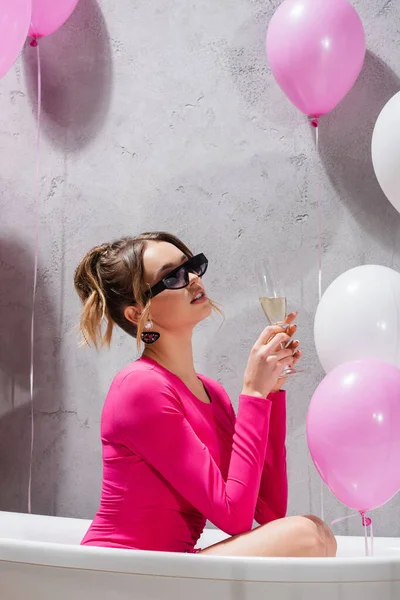 Stilvolle Frau hält Glas Champagner in Badewanne neben Luftballons — Stockfoto