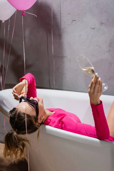Веселая женщина в солнечных очках держит шампанское возле воздушных шаров в ванной комнате — стоковое фото