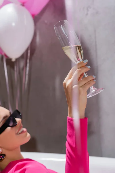 Copa de champán en mano de mujer sonriente en gafas de sol en bañera cerca de globos sobre fondo borroso - foto de stock