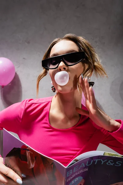 Visão de baixo ângulo de mulher elegante com bubblegum segurando revista perto de balão borrado — Fotografia de Stock