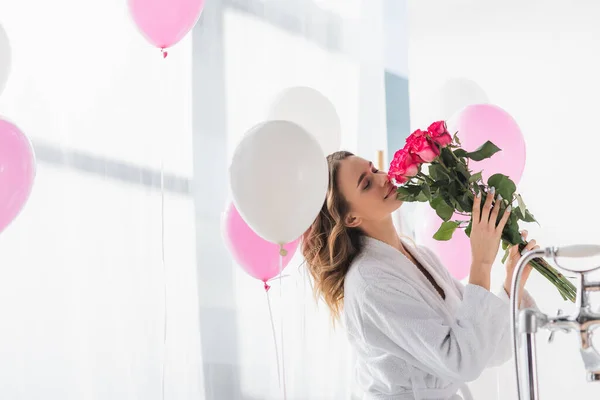 Jovem mulher em roupão de banho cheirando rosas perto de balões no banheiro — Fotografia de Stock