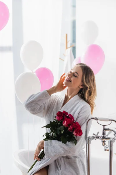 Mujer positiva en albornoz sosteniendo rosas cerca de la bañera y globos - foto de stock