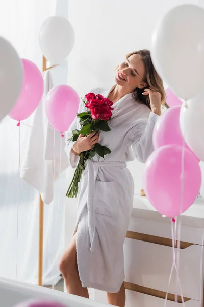 Positive woman in bathrobe holding roses near blurred balloons in bathroom — Stock Photo