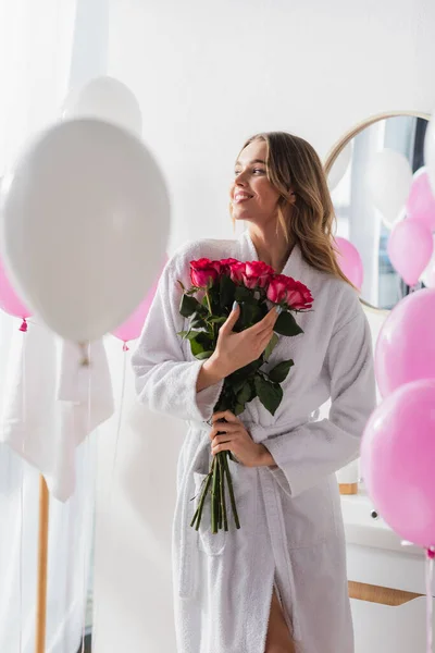 Young woman in bathrobe holding bouquet of roses near balloons in bathroom — Stock Photo