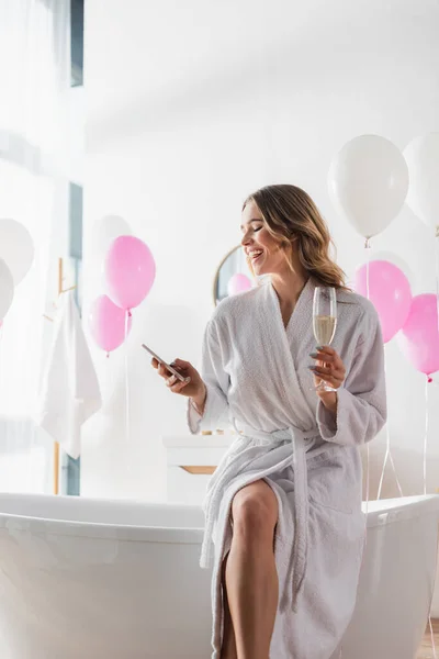 Smiling woman in bathrobe holding champagne and using smartphone near balloons in bathroom — Stock Photo