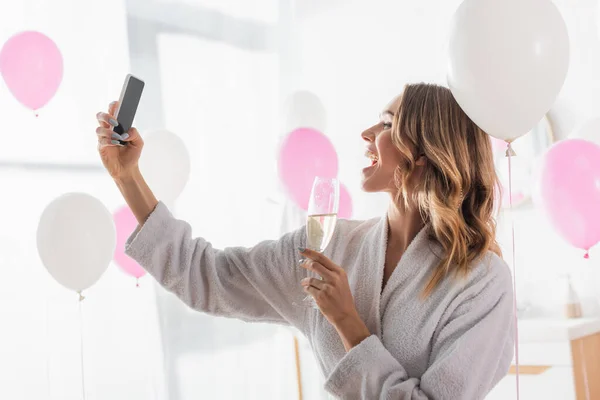 Vista laterale della donna allegra che prende selfie sullo smartphone e tiene lo champagne vicino ai palloncini — Foto stock