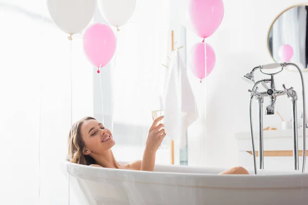 Smiling woman with champagne bathing near balloons in bathroom — Stock Photo