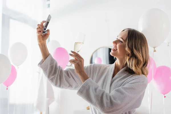 Mulher sorridente segurando champanhe e tirando selfie com smartphone no banheiro com balões — Fotografia de Stock