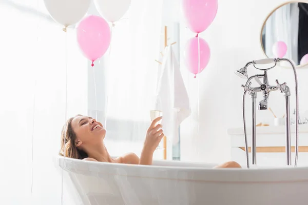 Happy woman holding glass of champagne near balloons in bathroom — Stock Photo