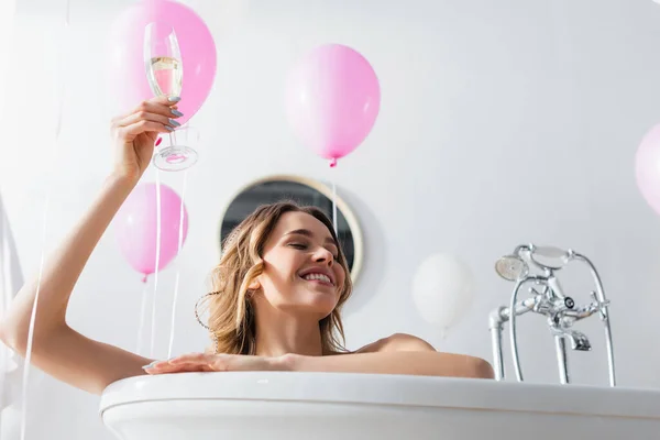 Vista de ángulo bajo de mujer sonriente con copa de champán sentada en bañera cerca de globos festivos - foto de stock