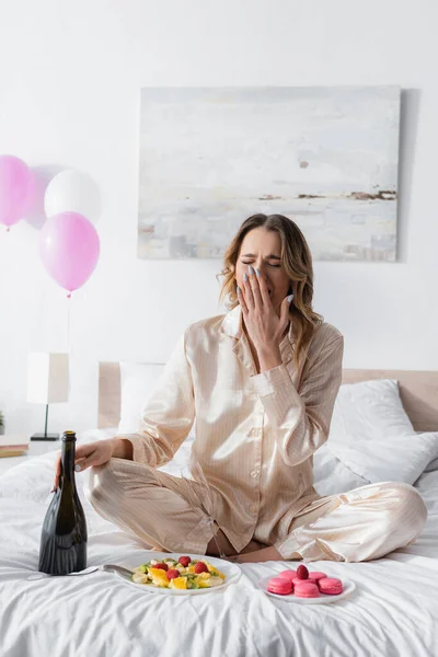 Woman with champagne yawning near fruit salad and macaroons on bed — Stock Photo
