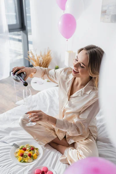 Mulher sorridente derramando champanhe perto de macaroons e salada de frutas na cama — Fotografia de Stock