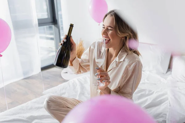 Mulher positiva segurando vidro e garrafa de champanhe na cama perto de balões festivos — Fotografia de Stock