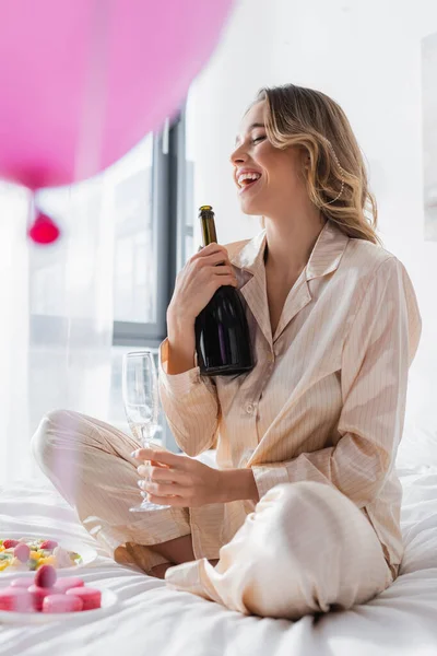 Happy woman with champagne sitting near macaroons, fruit salad and blurred balloon on bed — Stock Photo
