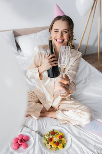 Femme en chapeau de fête tenant champagne et clin d'oeil à la caméra près de la salade de fruits et macarons sur le lit — Photo de stock