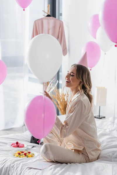 Positive woman holding champagne near balloons and food on bed — Stock Photo